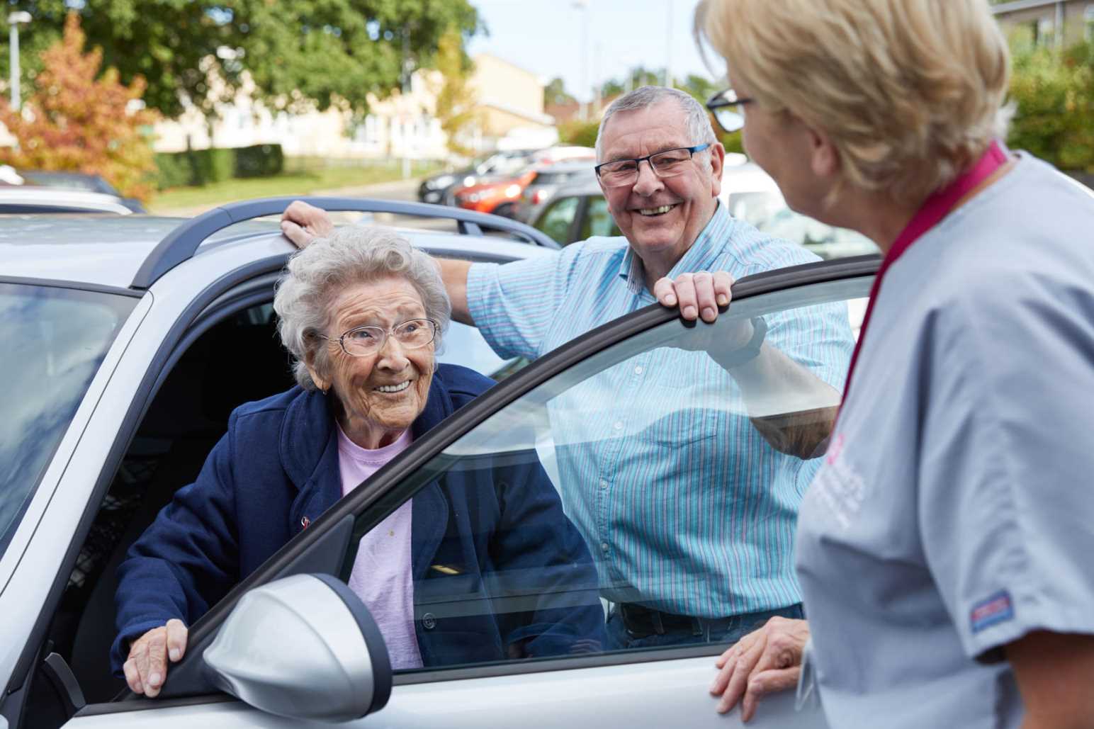 An elderly lady, smiling receiving help getting out of the car to visit our day hospice