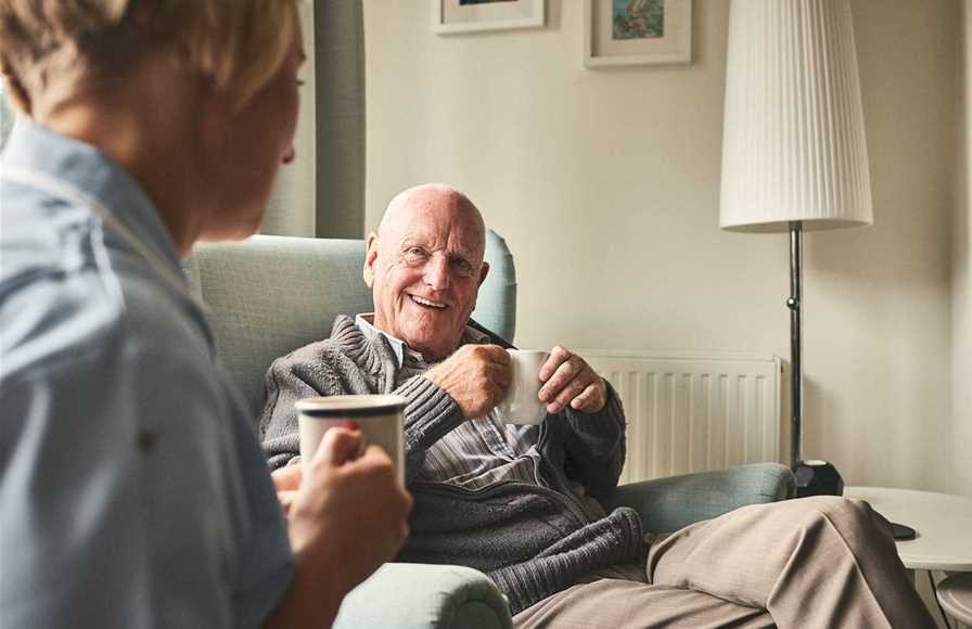 A man sitting in an arm chair in his home, chatting to a nurse